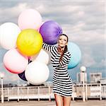 girl in black and white striped short dress and white high top sneakers sends air kiss holding bunch of multicolored balloons