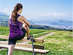Photo of a young woman stretching her leg before she starts to run on a country path.  City and lake in the distance.
