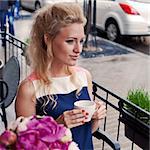 a beautiful young blond girl in summer dress at the table in pavement cafe holding a tea cup is looking thoughtfully in the distance