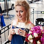 a beautiful young blond girl with blue eyes and toothy smile at the table in pavement cafe is holding a tea cup