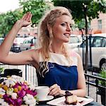 a beautiful young blond girl with a toothy smile in summer dress at the table in pavement cafe is waving someone