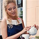 a beautiful young blond girl in summer dress at the table in pavement cafe is going to pour tea from the teapot into the cup