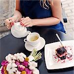 a beautiful young blond girl in summer dress at the table in pavement cafe is pouring green tea from the teapot into a white ceramic cup