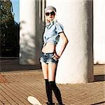 Young beautiful girl in checked knot shirt, stockings, jeans short shorts and sunglasses  stands with skateboard