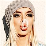 Portrait of beautiful young blond girl in beanie hat with her blown up bubble gum on her nose and plump lips