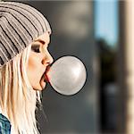 Portrait of beautiful young blonde teenage girl in beanie hat blows big bubble from bubble gum