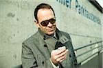 Close-up portrait of businessman wearing sunglasses and overcoat, looking at cell phone outdoors, Germany