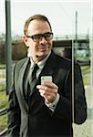 Portrait of businessman wearing glasses and holding cell phone, standing at train station outdoors, Mannheim, Germany