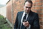 Portrait of businessman standing next to brick wall outdoors, looking at cell phone, Germany