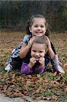 Two sisters tumble in the grass and leaves on a Fall morning.  One sister sits on the other one and they both are smiling and giggling together.