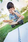 Frustrated Cute Young Boy Holding Pencil Sitting on the Grass Outdoors.