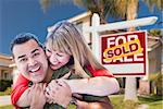 Happy Hugging Mixed Race Couple in Front of Sold Home For Sale Real Estate Sign and House.