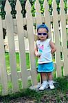 Little girl wearing sunglasses and a cap turned backwards, stands besides a wooden picket fence.