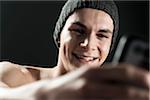 Close-up portrait of young man wearing toque, taking a selfie with cell phone, studio shot on black background