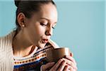 Close-up of teenage girl holding mug and blowing on hot drink, studio shot on blue background