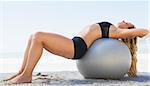 Fit blonde stretching her back on exercise ball at the beach on a sunny day