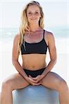 Fit blonde sitting on exercise ball at the beach smiling at camera on a sunny day