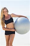 Fit blonde holding exercise ball at the beach smiling at camera on a sunny day