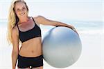 Fit blonde holding exercise ball at the beach smiling at camera on a sunny day