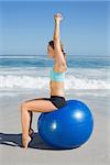 Fit woman sitting on exercise ball at the beach stretching arms on a sunny day