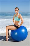 Fit woman sitting on exercise ball at the beach smiling at camera on a sunny day