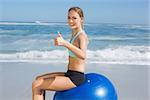 Fit woman sitting on exercise ball at the beach smiling at camera on a sunny day