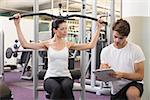 Fit brunette using weights machine for arms with trainer taking notes at the gym