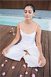 Peaceful brunette in white sitting in lotus pose surrounded by petals in health spa