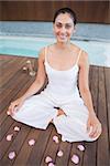 Peaceful brunette in white sitting in lotus pose surrounded by petals in health spa