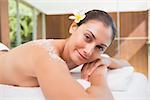 Beautiful smiling brunette lying on massage table with salt scrub on back in the health spa