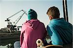 Backview of teenage girl and boy sitting on bench outdoors with skateboard, industrial area, Mannheim, Germany