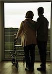 Grandmother using walker standing beside teenage grandson, and looking out of window, Germany