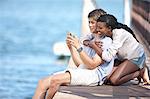 Young couple sitting on jetty, looking at mobile phone