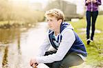 Unhappy teenage boy crouching on riverbank