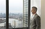 Portrait of mature businessman looking out of office window, Canary Wharf, London, UK