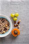 A bowl of muesli next to grapes, nuts and an apricot