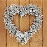 Heart shaped christmas wreath with silver thistle, bow and bauble decorations over old oak background.