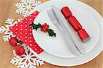 Christmas dinner place setting with white plates, red bauble decorations,cutlery, snowflakes and holly over oak background.
