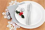 Christmas dinner place setting with white plates, cutlery, name tag, holly, silver bauble and snowflake decorations over oak table background.