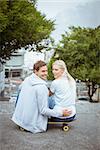 Hip young couple sitting on skateboard smiling at camera on a sunny day in the city