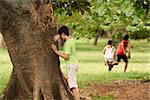 young boys and girls playing hide and seek in park, with kid counting leaning on tree