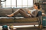 Happy young woman laying on divan and using tablet pc in loft apartment