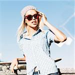 Close-up of smiling blond girl in beanie hat and leopard sunglasses who leans on skateboard adjusting her hair on sunny day in the street