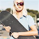 Close-up of beautiful  blond girl in beanie hat and leopard sunglasses who hugs skateboard on sunny day in the street