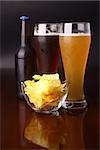 Two glasses of beer, bottle and potato chips over a dark background