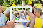 Hand holding tablet pc showing extended family wearing party hats at birthday celebration in park