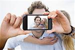Hand holding smartphone showing man piggybacking woman at beach