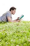 Side view of man reading book while lying on grass against clear sky