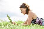Side view of young woman using Tablet PC while lying on grass against sky