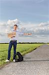 Full length of young man hitching while holding anywhere sign on countryside